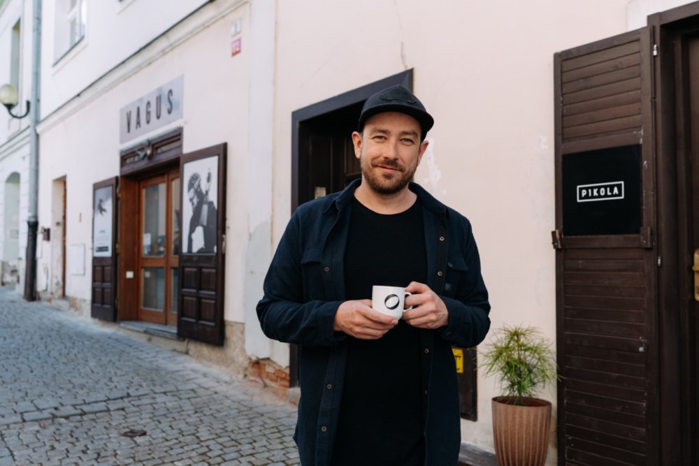 Váša Láznička stojí se šálkem kávy v ruce před kavárnou Pikola a v pozadí je vidět i Vagus