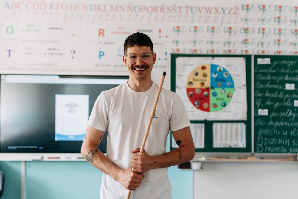 Matěj Feranc stojí před tabulí ve třídě, usmívá se a v rukách drží ukazovátko.