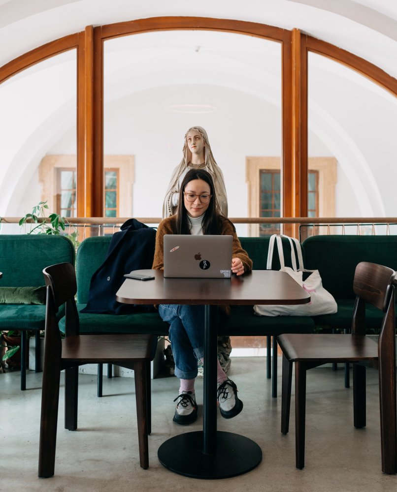 Tereza Dehnerová sedí v kavárně u dřevěného stolu, má před sebou počítat Apple Macbook a za ní je vidět socha Panny Marie